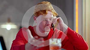 Portrait of irritated young redhead Caucasian man talking on the phone sitting at bar counter in pub. Stressed
