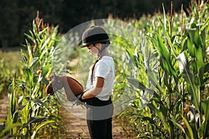 Portrait of a jockey girl in a helmet and a white t-shirt, who sits on a brown toy horse in a corn field photo