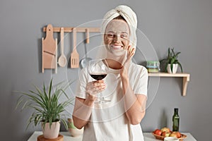 Portrait of joyful adorable woman with cosmetic mask in white towel wrapped around head drinking wine in kitchen, enjoying her