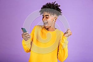 Portrait of joyful african american man smiling and holding smartphone, while listening to music via bluetooth earpod, isolated o