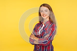 Portrait of joyful attractive ginger girl in checkered shirt standing with crossed hands and smiling charmingly at camera
