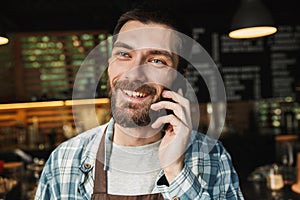 Portrait of joyful barista guy talking on cellphone in street cafe or coffeehouse outdoor