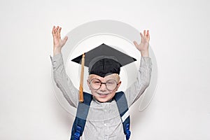 Portrait of a joyful blond boy in big glasses, academic hat and a backpack. Hands up. White background