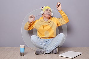 Portrait of joyful cheerful happy man wearing beanie hat, yellow hoodie and jeans posing against gray wall, listening musing with