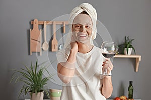 Portrait of joyful cheerful woman with cosmetic mask in white towel wrapped around head drinking wine in kitchen, keeps hand on