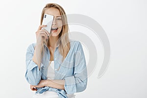 Portrait of joyful confident european female with fair hair, covering half of face with brand new smarpthone, holding