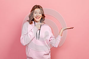 Portrait of joyful curly haired teenage girl in hoodie pointing finger right at empty copy space with toothy smile, place for