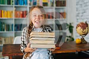 Portrait of joyful elementary child school girl holding stack of books in library at school, looking at camera.