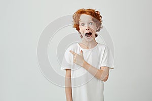 Portrait of joyful ginger child with freckles posing with opened mouth and crazy expression, pointing at free space for