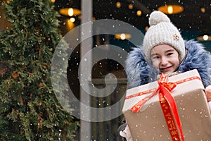 Portrait of joyful girl with a gift box for Christmas on a city street in winter with snow on a festive market with decorations