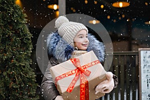 Portrait of joyful girl with a gift box for Christmas on a city street in winter with snow on a festive market with decorations