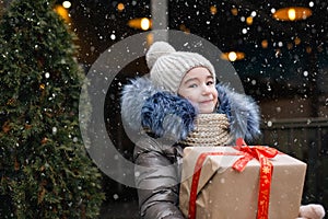 Portrait of joyful girl with a gift box for Christmas on a city street in winter with snow on a festive market with decorations