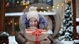 Portrait of joyful girl with a gift box for Christmas on a city street in winter with snow on a festive market with decorations an