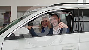 portrait of joyful man with his wife sitting at wheel of a car and showing keys to a new car from auto center, smiling