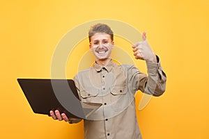 Portrait of joyful nerd in glasses and shirt with laptop in hand on yellow background, looks into camera and shows thumbs up.