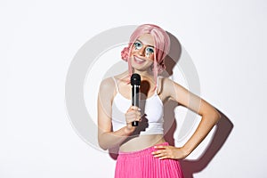 Portrait of joyful party girl in pink wig, bright halloween makeup, singing in microphone, standing over white