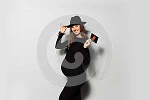 Portrait of joyful pregnant woman, wearing black dress and hat, holding ultrasound scan of her baby.