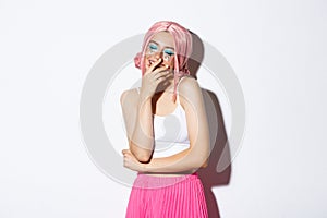 Portrait of joyful pretty girl in pink wig, dressed for halloween party, laughing and smiling, standing over white