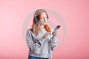 Portrait of a joyful redhead girl who listens to music on headphones with her smartphone in the studio on a pink background.