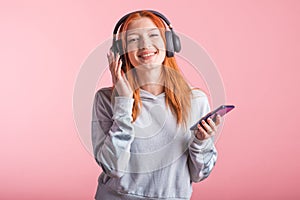 Portrait of a joyful redhead girl who listens to music on headphones with her smartphone in the studio on a pink background.