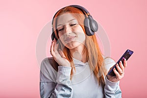 Portrait of a joyful redhead girl who listens to music on headphones with her smartphone in the studio on a pink background.