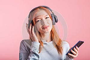 Portrait of a joyful redhead girl who listens to music on headphones with her smartphone in the studio on a pink background.