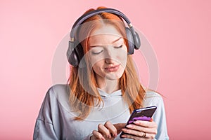 Portrait of a joyful redhead girl who listens to music on headphones with her smartphone in the studio on a pink background.