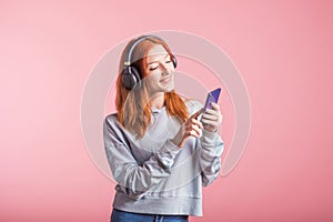 Portrait of a joyful redhead girl who listens to music on headphones with her smartphone in the studio on a pink background.