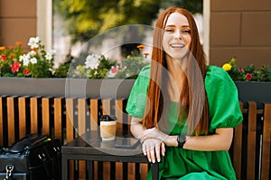 Portrait of joyful redhead young woman sitting at table with coffee cup and mobile phone in outdoor cafe terrace in