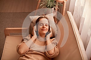 Portrait of joyful relaxed caucasian young woman listening music while resting on couch at home, keeps eyes closed, being calm and