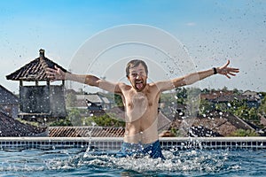 Portrait of joyful slender man with chest hair, emerges from pool, water splashes, shows tongue and hands up, on hotel roof.