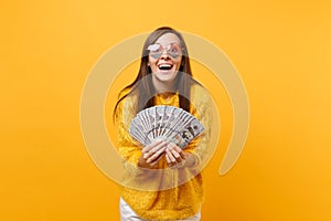 Portrait of joyful smiling young woman in heart eyeglasses holding bundle lots of dollars cash money isolated on bright