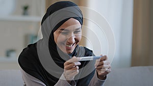 Portrait joyful surprised girl in hijab holding pregnancy test sit in living room excited with positive result beautiful