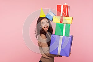 Portrait of joyful surprised young woman wearing party cone and looking with amazement at many gift boxes in her hands