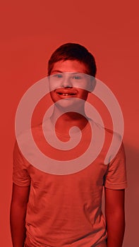 Portrait of joyful teenaged disabled boy with Down syndrome smiling at camera while posing isolated over red light