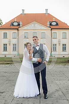 Portrait of joyful wedding couple standing outside near old building in summer. Young gorgeous woman hugging young man.