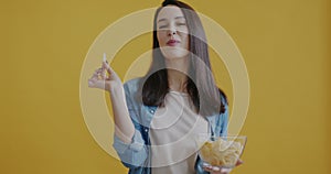 Portrait of joyful woman eating potato chips with pleasure smiling on yellow color background