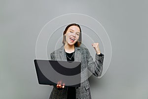 Portrait of joyful woman in smart casual clothes and laptop in hand rejoices with raised hand on gray background, looks at camera