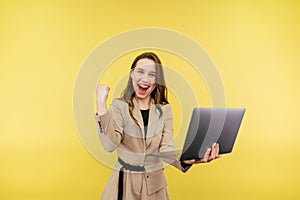 Portrait of a joyful woman in a suit on a yellow background, holding a laptop and emotionally happy with shouts and with raised