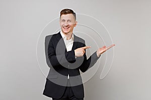 Portrait of joyful young business man in classic black suit pointing index finger hand aside isolated on grey wall
