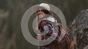 portrait of joyful young female hiker in hat standing on cliff on forest background and looking with smile, mountain