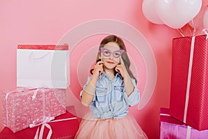 Portrait joyful young girl in blue shirt holding mask on face suround colorful giftboxes on pink background. Lovely