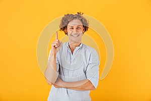 Portrait of joyful young man 20s smiling and pointing finger upward at copyspace, isolated over yellow background