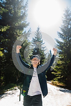 Portrait of a joyful young man in casual clothes and a shirt while walking in the mountains in the woods with arms raised and