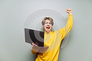 Portrait of a joyful young man in casual clothes stands with a laptop in his hands on a background of gray wall and rejoices in