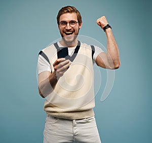 Portrait of a joyful young man holding mobile phone isolated over blue background, celebrating.