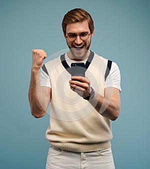 Portrait of a joyful young man holding mobile phone isolated over blue background, celebrating.
