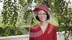 Portrait of joyful young woman graduating student in gown and mortar-board smiling and looking at camera standing under