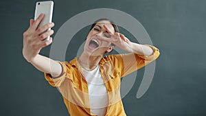 Portrait of joyful young woman taking selfie with smartphone camera showing hand gestures