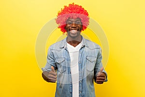 Portrait of joyous funny carefree man in red curly wig, showing thumbs up. indoor studio shot isolated on yellow background